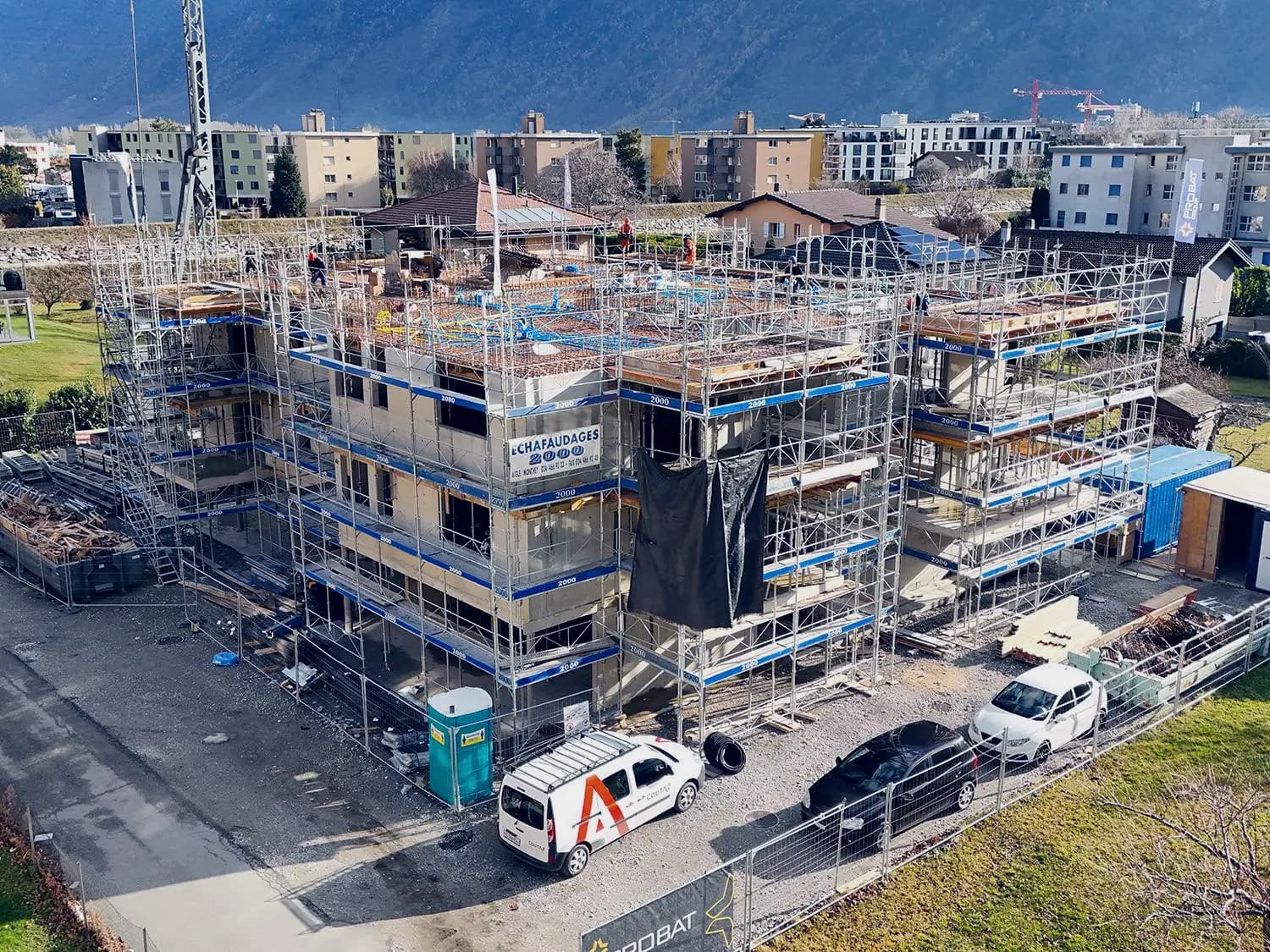 Immeuble résidentiel en construction, entouré d’échafaudages, dans la plaine ; la dalle du dernier étage est en cours de ferraillage, montagnes à l’arrière-plan